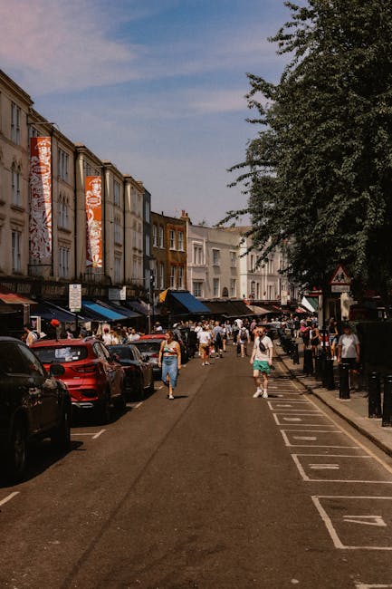 A busy street scene in Notting Hill during daytime, showing pedestrians walking along the pavement next to parked cars and closely lined buildings with multiple stories and shopfronts. Several large c