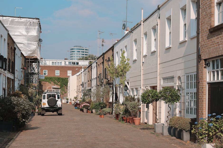 A busy street scene in Notting Hill during daytime, showing pedestrians walking along the pavement next to parked cars and closely lined buildings with multiple stories and shopfronts. Several large c
