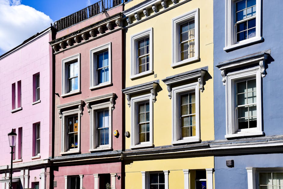 The image shows the facades of three colourful terraced houses in a city neighbourhood, each with distinctive pastel colors: pink on the left, yellow in the center, and blue on the right. The building