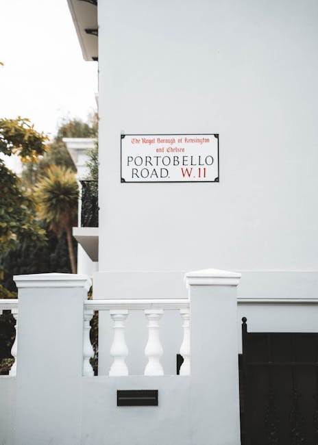 A close-up view of a white residential building with a white balustrade and a black mailbox, situated on Portobello Road in Notting Hill. The exterior wall is smooth with a street sign indicating 'Portobello Road W.11', along with an additional sign referencing the Royal Borough of Kensington and Chelsea. The image captures part of the doorway and the lower section of the building, with a backdrop of blurred trees and foliage in the distance. This setting reflects a typical property where house removals and furniture transport services by Notting Hill Man and Van might occur as part of a home relocation process. The scene emphasizes the building's exterior entrance, which could be relevant for loading or unloading furniture during a move, with natural lighting suggesting daytime conditions.