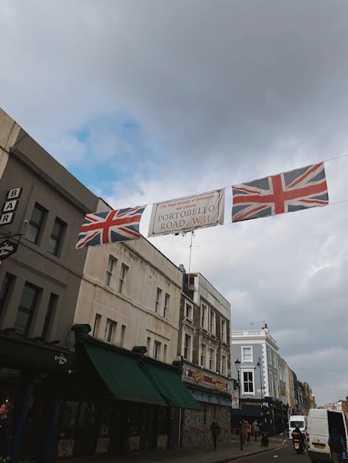 The image shows a daytime street scene in Notting Hill with a banner hanging above the pavement, decorated with two Union Jack flags on either side and a central sign reading 'Portobello Road W11'. Below the banner, a group of movers from Notting Hill Man and Van are engaged in furniture transport and home relocation activities. They are loading cardboard boxes, some wrapped in plastic, onto a large black van parked along the street. The loading process involves a green trolley and the movers are wearing casual work clothing. Surrounding buildings include shops with awnings and residential properties, under a cloudy sky. The scene captures the logistics and packing involved in a house removal or moving service within the neighbourhood, highlighting the detailed actions of loading and transport preparation.