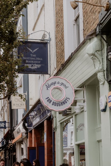 A close-up view of a white residential building with a white balustrade and a black mailbox, situated on Portobello Road in Notting Hill. The exterior wall is smooth with a street sign indicating 'Portobello Road W.11', along with an additional sign referencing the Royal Borough of Kensington and Chelsea. The image captures part of the doorway and the lower section of the building, with a backdrop of blurred trees and foliage in the distance. This setting reflects a typical property where house removals and furniture transport services by Notting Hill Man and Van might occur as part of a home relocation process. The scene emphasizes the building's exterior entrance, which could be relevant for loading or unloading furniture during a move, with natural lighting suggesting daytime conditions.