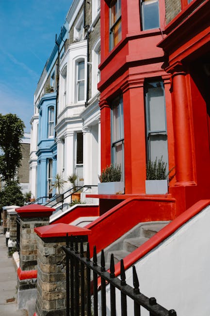 A row of Victorian-style terraced houses with colourful facades, including a prominent red-painted building with decorative columns and large windows, set along a tree-lined street under a clear blue