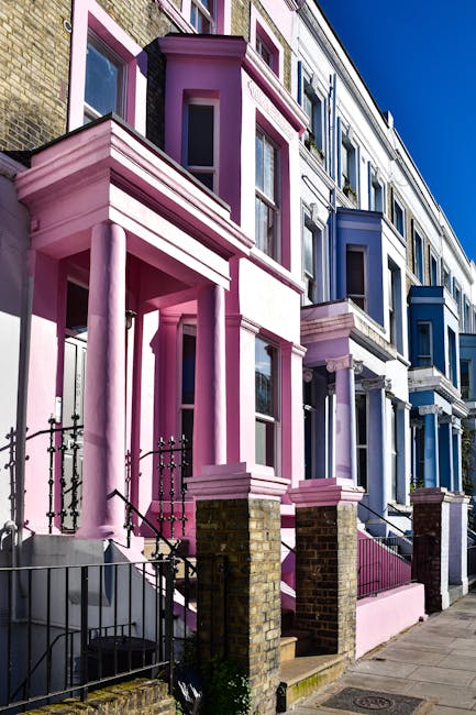 A row of Victorian-style terraced houses with colourful facades, including a prominent red-painted building with decorative columns and large windows, set along a tree-lined street under a clear blue