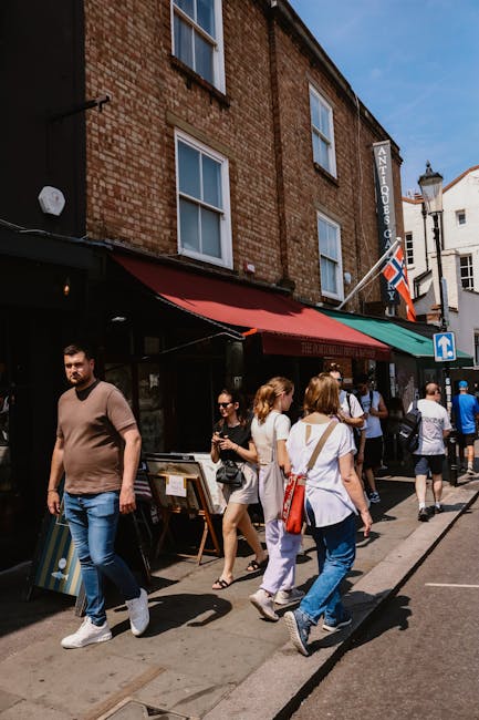 A busy street scene outside a brick building with a red and green striped awning, on the facade of which is a sign reading 'ANTIQUES'. Several pedestrians, including men and women dressed casually, wa