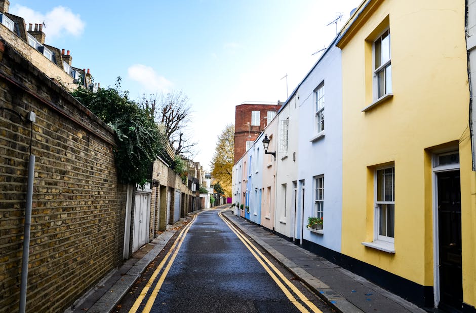 A narrow residential street in Notting Hill with a slight curve, featuring a yellow-painted house with white-framed sash windows on the right side and a brick wall with garages and greenery on the left. The street is paved with tarmac, showing a few wet patches, and has double yellow lines along the kerb. In the background, a red brick building rises above the row of houses, and trees with autumn foliage are visible under a partly cloudy sky. This scene captures the typical setting for house removals and furniture transport, with the street's tight width indicating potential logistical challenges in moving larger items, which Notting Hill Man and Van may assist with during home relocation and packing and moving services.