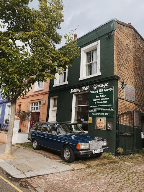The image shows a two-storey building with a dark green exterior and white window frames, identified as Notting Hill Garage. A large green sign mounted on the facade displays text indicating services such as sales, repairs, and classic car care, along with a contact phone number. Parked directly in front of the garage is a vintage blue station wagon with a roof rack, positioned on a paved sidewalk adjacent to a residential street. A small tree with green foliage is situated to the left, partially obscuring the lower part of the building and casting some shadows on the pavement. The surrounding area includes a neighboring terraced house with a peach-colored facade and white windows. The street appears quiet with no other vehicles or pedestrians visible, and the overall scene is illuminated by natural daylight with an overcast sky. This setting is typical of a London neighbourhood where local garages and residential buildings coexist, often involved in home relocation and furniture transport services, which [COMPANY_NAME] provides as part of their removals offerings. The image captures the loading area outside the garage, highlighting the urban environment suitable for house removals and moving logistics in Notting Hill.
