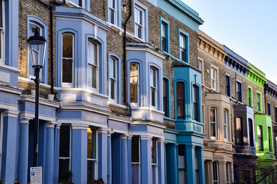 A row of white terraced houses in Notting Hill with multiple stories, tall windows, and decorative mouldings, featuring small balconies with wrought iron railings. In front of the buildings, there is 